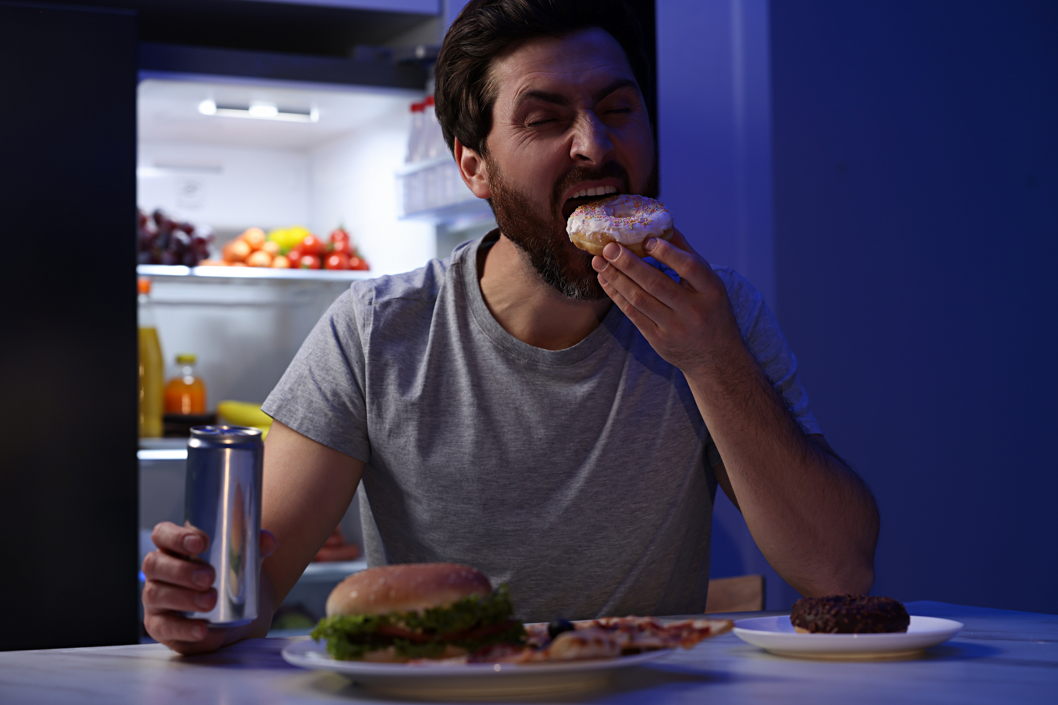 Man holding can and eating donut in kitchen at night. Bad habit